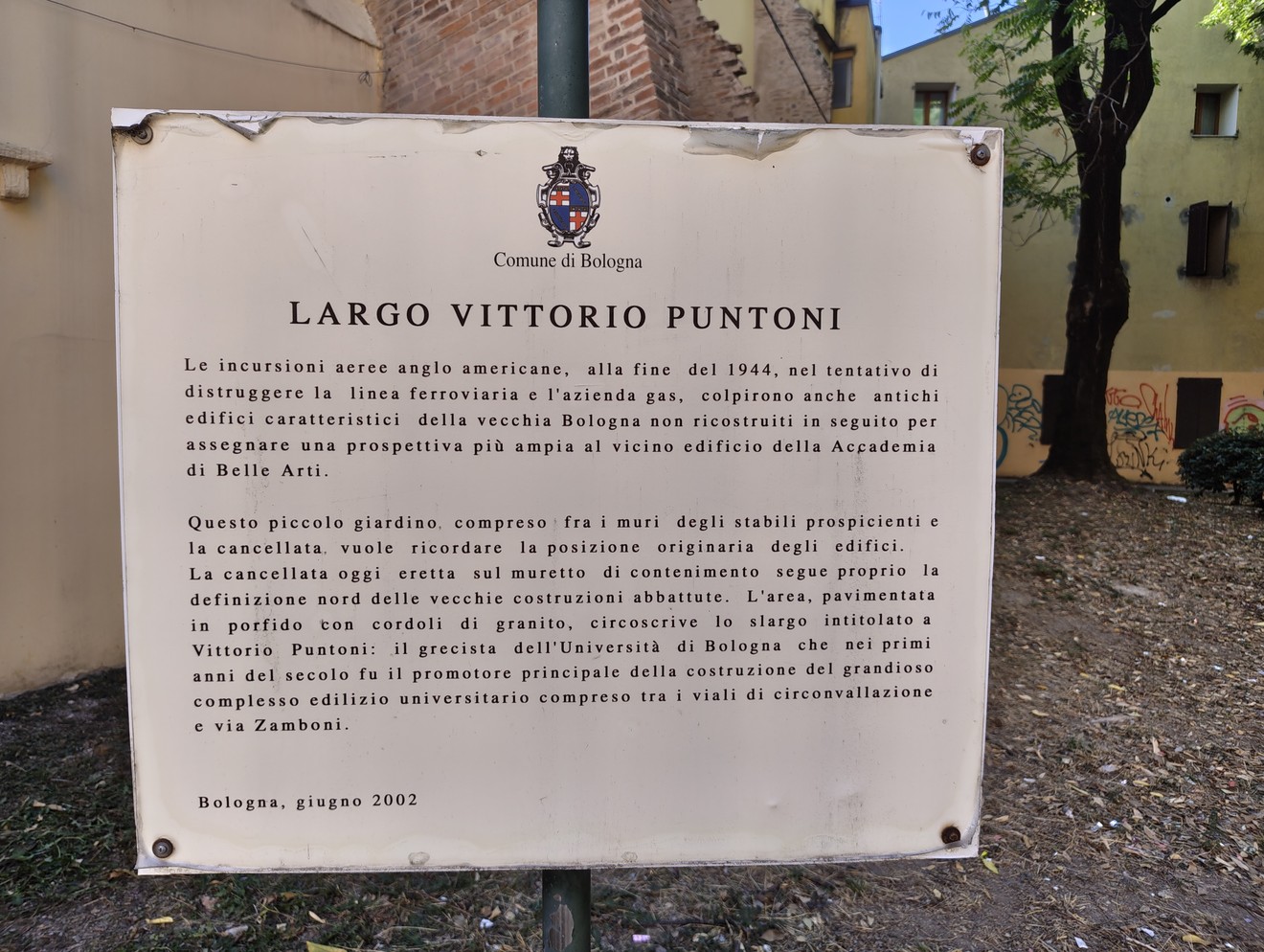 Targa commemorativa in metallo con stemma e iscrizione, fissata su parete esterna in un cortile.