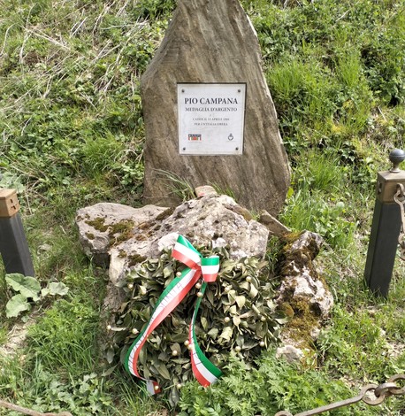 Cippo commemorativo di Pio Campana con alloro e nastri tricolore, immerso nel verde della natura.