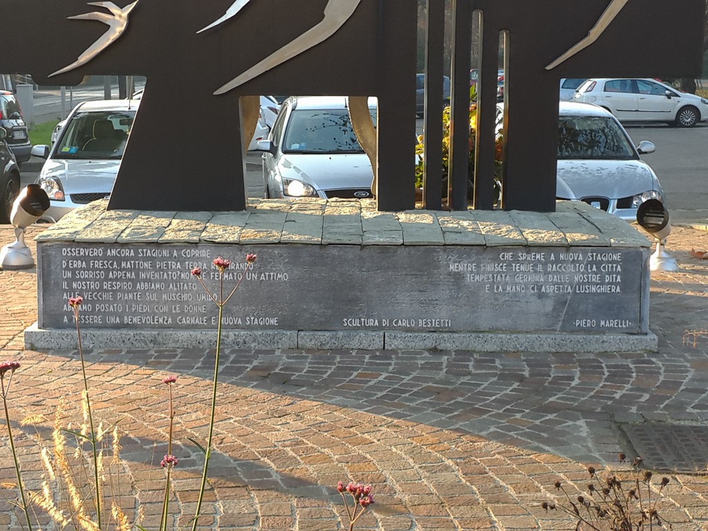 Monumento in pietra con iscrizione, sormontato da sagome metalliche di rondini, su una piazza con auto parcheggiate.