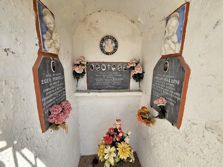 Nicchia bianca con due lapidi ai lati, una targa centrale e fiori artificiali, dipinti di Madonna sopra le lapidi.