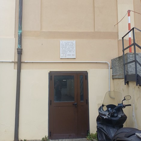 Lapide con iscrizione su parete esterna, sopra una porta, tra una grondaia e una scala antincendio.