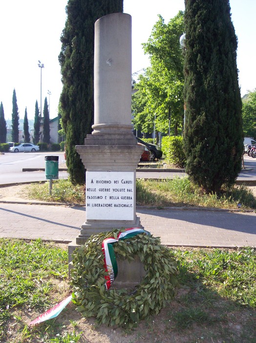 Cippo commemorativo dedicato ai caduti, adornato da una corona di alloro e circondato da alberi.