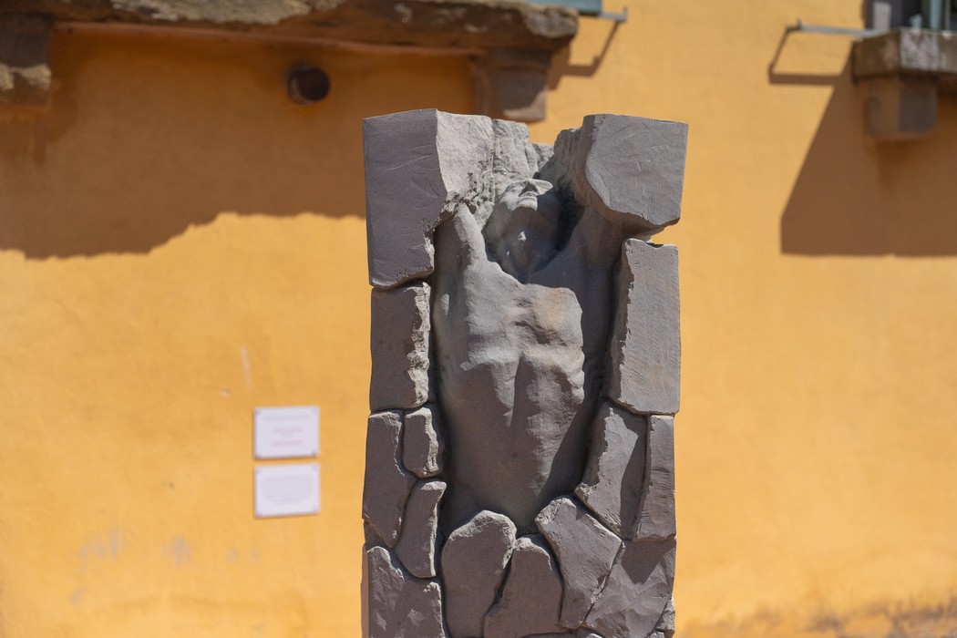 Statua in pietra scolpita a forma di figura umana emergente da un blocco squadrato, davanti a una parete gialla.