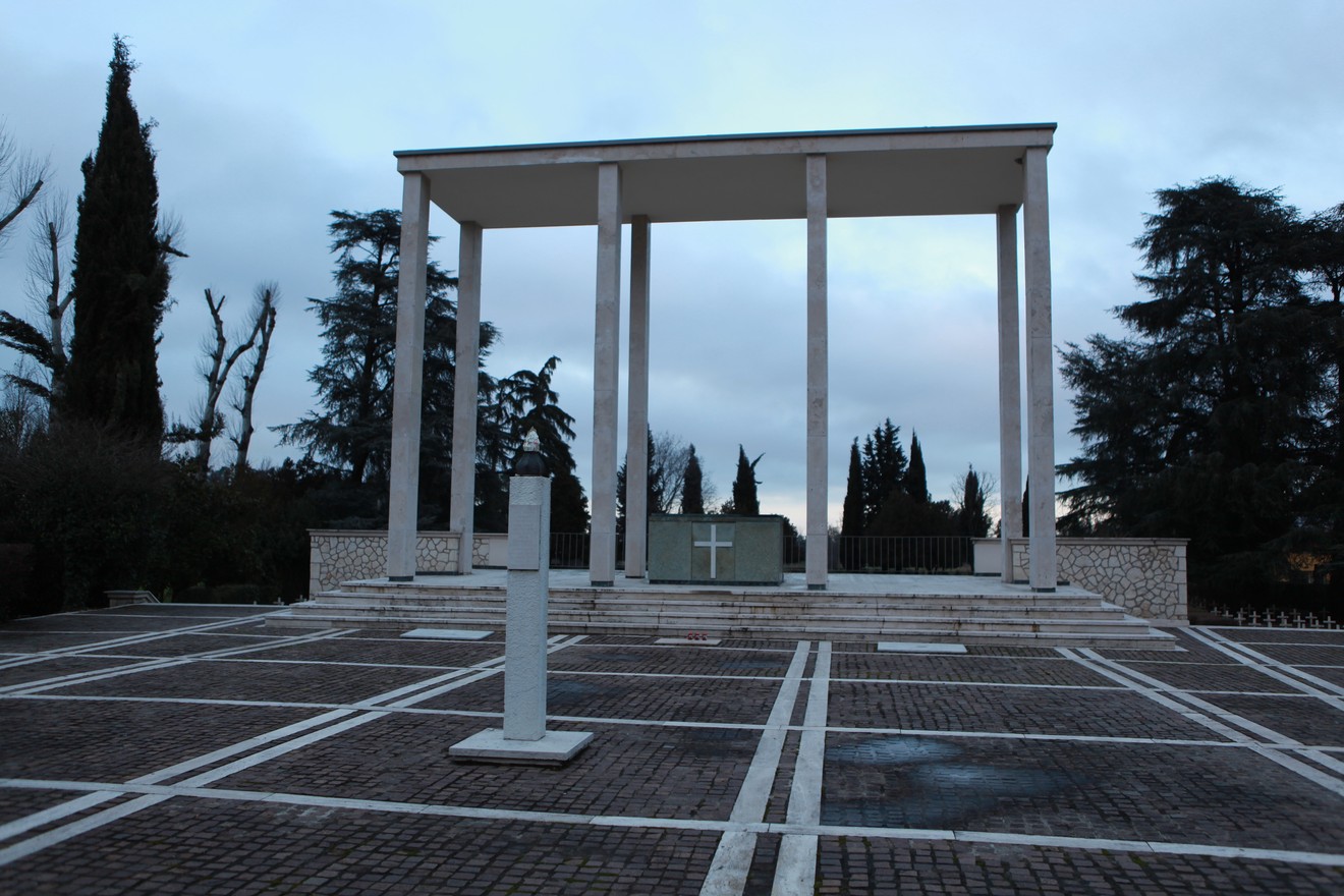 Cimitero di guerra polacco con monumento architettonico e alberi circostanti sotto un cielo nuvoloso.