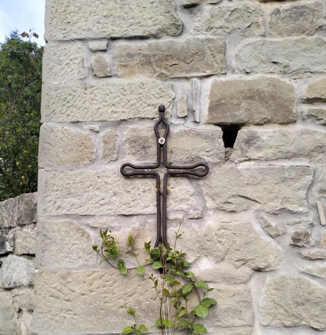 Croce in metallo montata su un muro di pietra, con piante verdi che crescono vicino.
