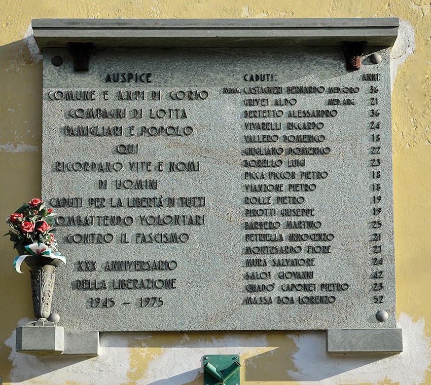 Lapide in pietra su parete, con iscrizione e doppio elenco di nomi; sotto, un vaso con fiori.