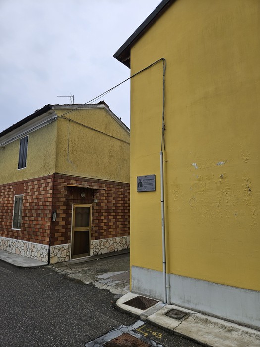 Targa commemorativa su parete gialla di edificio, tra due case in un angolo di strada.