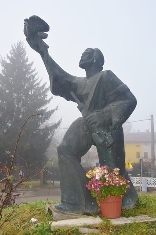 Monumento al Partigiano con figura eretta che regge un berretto, circondata da fiori in un contesto nebbioso.