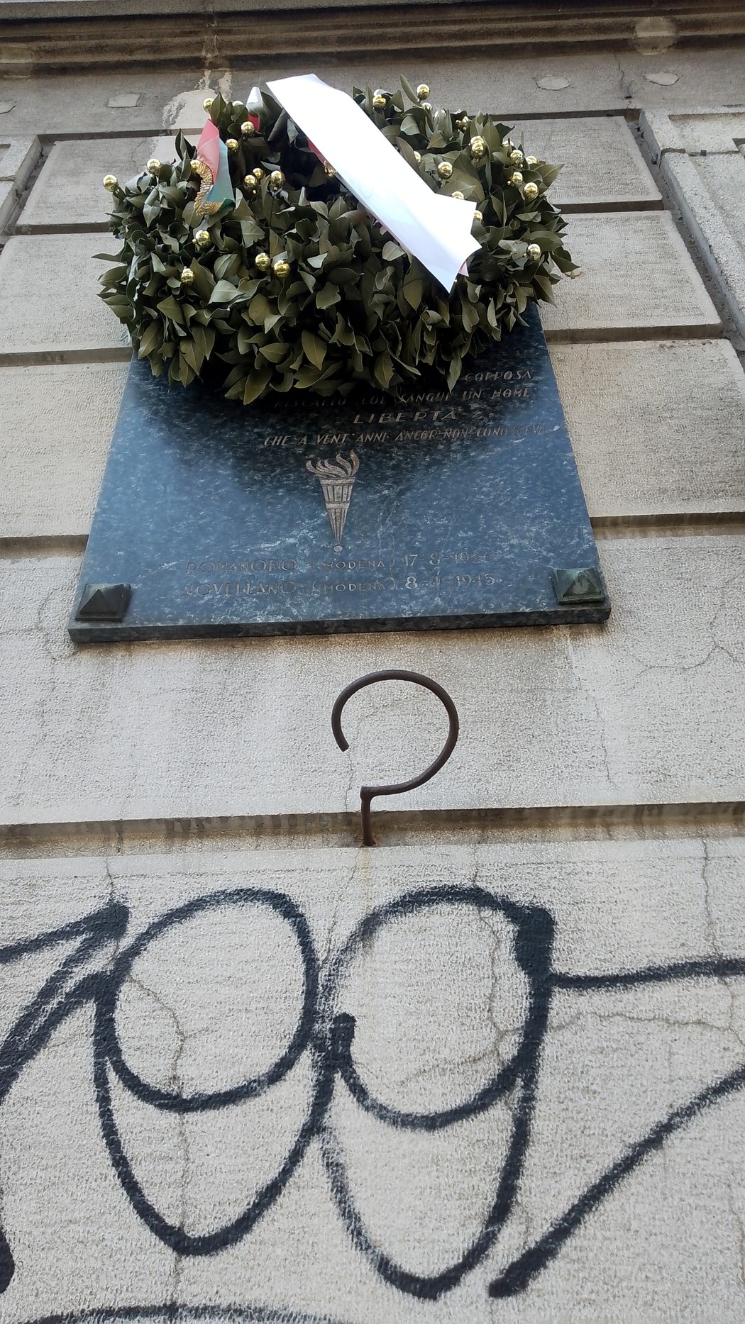 Lapide commemorativa di Rinaldi con ghirlanda di fiori, affissa su una parete con graffiti.