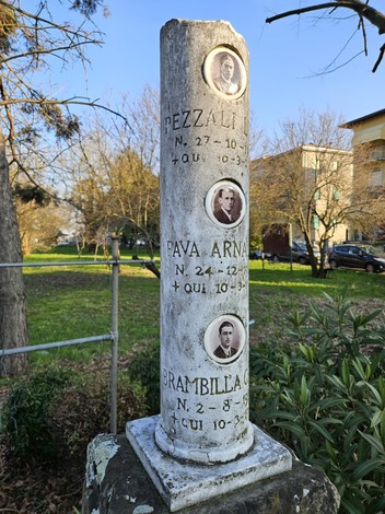 Cippo commemorativo con ritratti di Brambilla, Fava e Pezzali in un contesto verde e urbano.