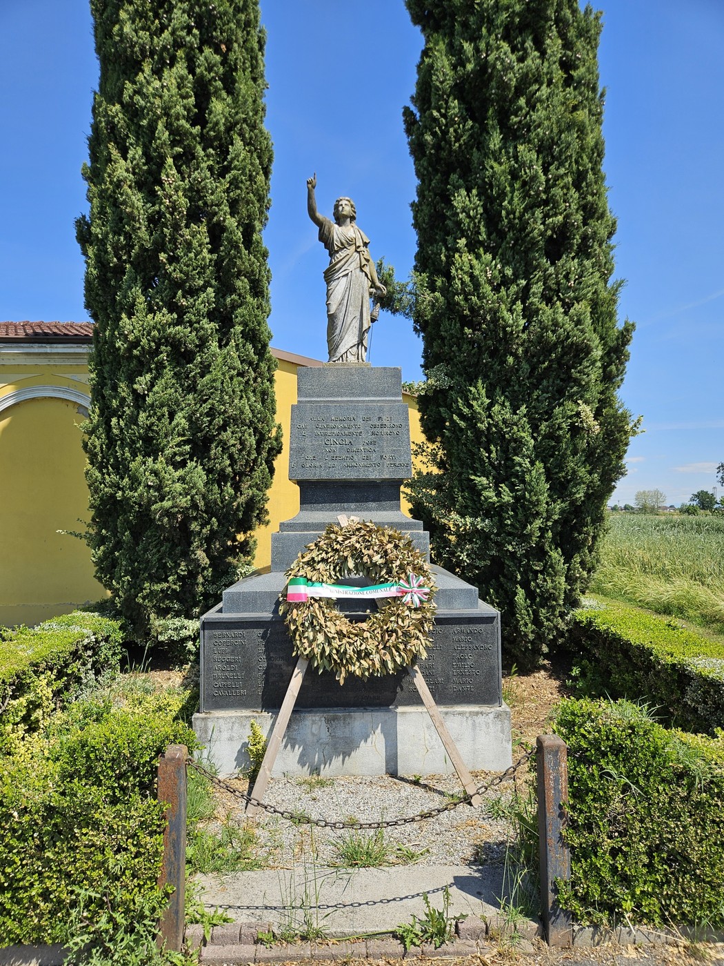 Monumento ai Caduti di Cingia de' Botti, con statue e corona d'alloro, circondato da piante e verde.