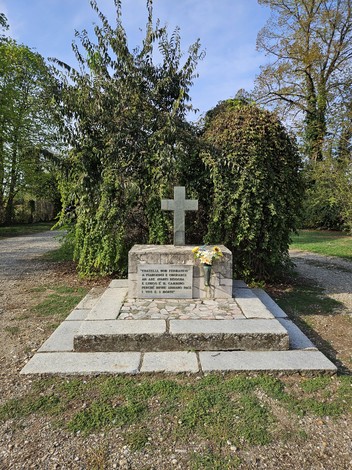 Monumento in pietra con croce, lapide con iscrizione e fiori, circondato da vegetazione in un parco.