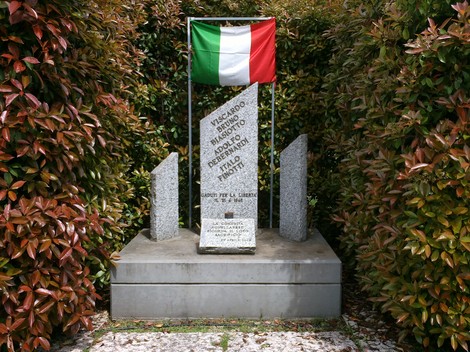 Cippo commemorativo dedicato a De Bernardi, Biasotto e Finotti, circondato da vegetazione e con bandiera italiana.