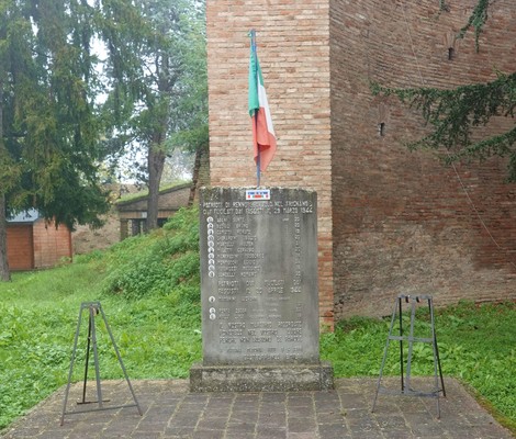 Monumento commemorativo con epigrafi, bandiera italiana e sfondo di un forte militare.