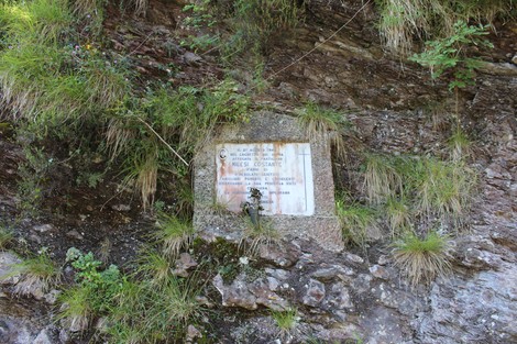 Lapide commemorativa affissa su una roccia, circondata da vegetazione e piante.