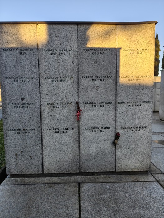 Monumento commemorativo al "Campo della Gloria", con iscrizioni e ombre sui pannelli. Atmosfera di rispetto e memoria.