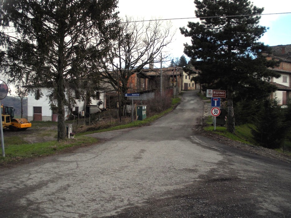 Strada in direzione di Ruino, con un paesaggio tipico montano e case storiche visibili sullo sfondo.