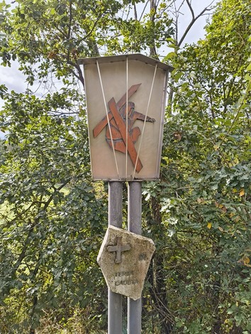 Pannello della Via Crucis di Casaglia, decorato con simboli religiosi e circondato da vegetazione.