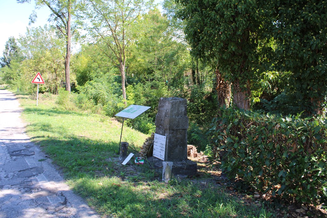 Cippo in pietra con iscrizione e targa ai bordi di una strada sterrata, tra vegetazione.