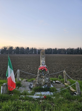 Cippo commemorativo con fiori, bandiera italiana e paesaggio rurale sullo sfondo.
