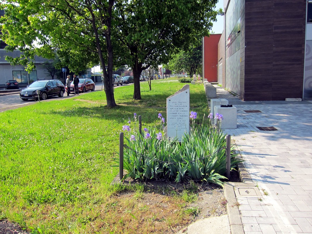 Cippo commemorativo circondato da fiori in un'area verde, con alberi e un marciapiede sullo sfondo.