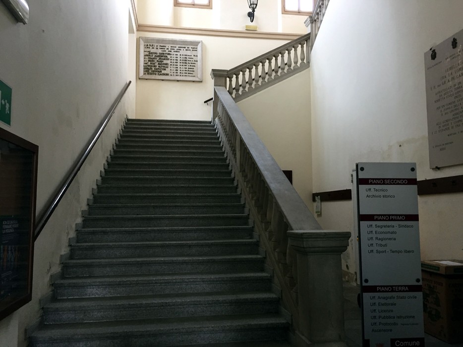 Scalinata all'interno di un edificio, con una lapide commemorativa sulla parete e segnaletica direzionale visibile.