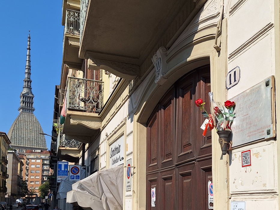 Targa su parete di palazzo, accanto a un portone ligneo, con fiori e iscrizione sottostante.