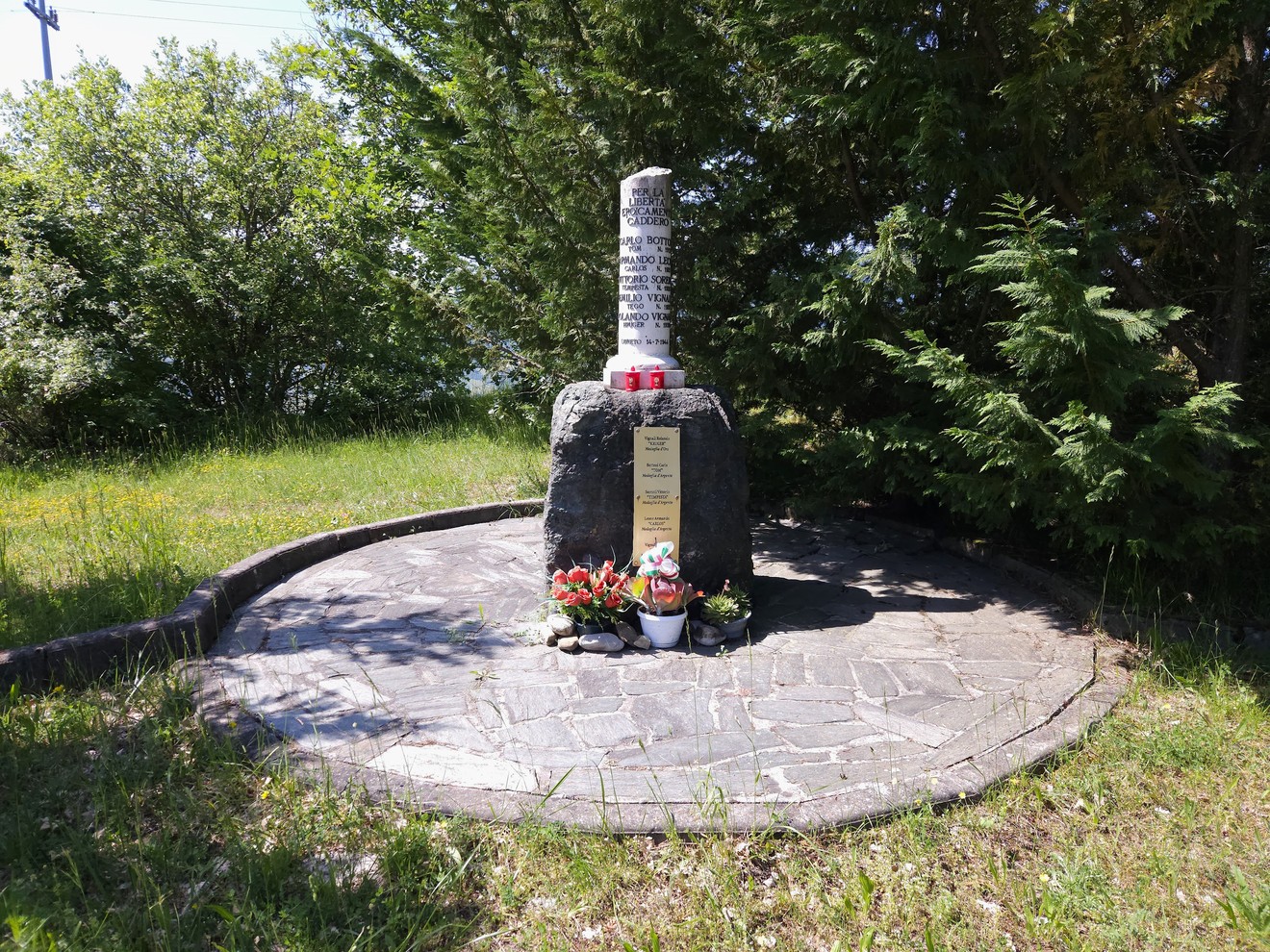 Cippo in pietra con iscrizione su base di roccia, fiori ai piedi, area pavimentata circolare tra alberi.