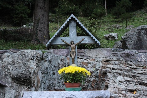 Monumento commemorativo con croce e statua di Cristo, circondato da fiori e un paesaggio naturale.
