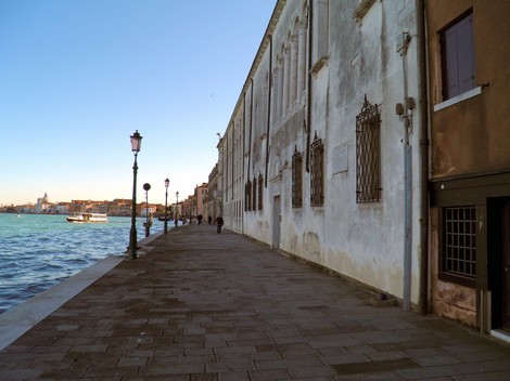 Marciapiede lungo il canale, con edifici storici e lampioni, sullo sfondo una barca e il sole che tramonta.