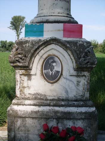 Cippo commemorativo con foto e decorazioni tricolori, immerso nella natura.