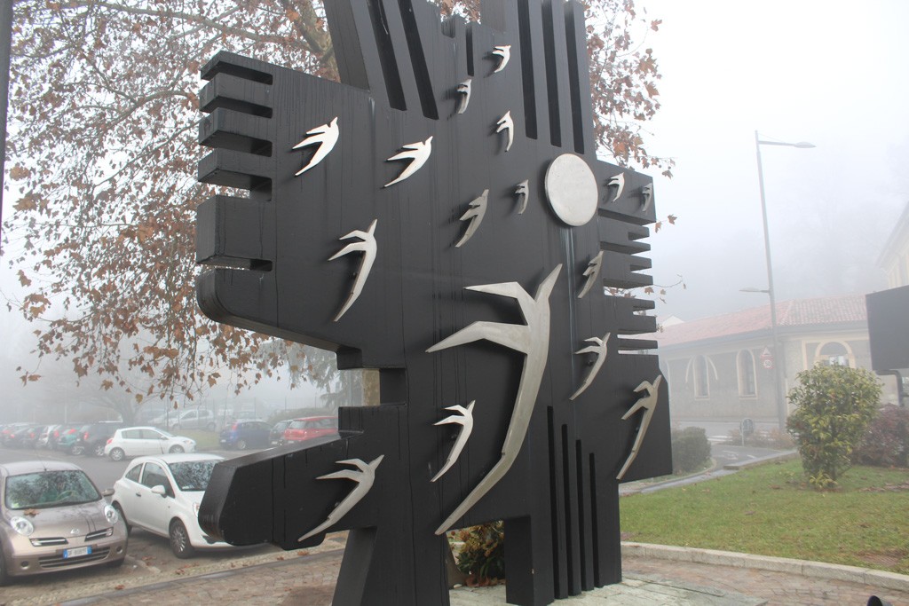 Monumento ai Caduti in metallo scuro con sagome di rondini stilizzate, collocato in una piazza.