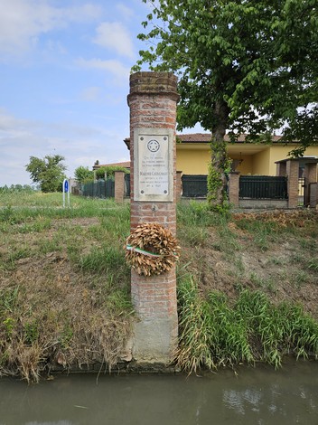Cippo di commemorazione a Cavagnari, con corona d'alloro e vegetazione circostante lungo un canale.
