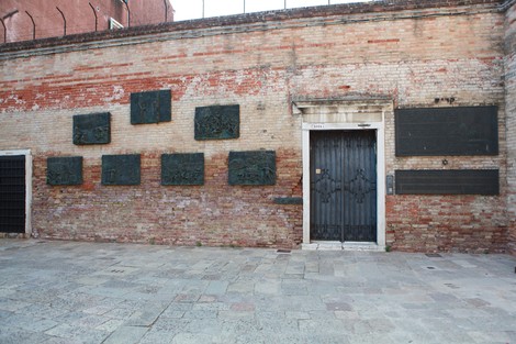 Monumento all'Olocausto con pannelli in bronzo su muro di mattoni e una porta centrale, in un contesto urbano.