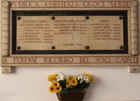 Lapide commemorativa ai caduti della Croce Verde, con fiori gialli in primo piano, in un ambiente sobrio e rispettoso.