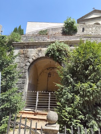Cappella in pietra con arco a tutto sesto e cancellata metallica, iscrizione sull'architrave, circondata da alberi.