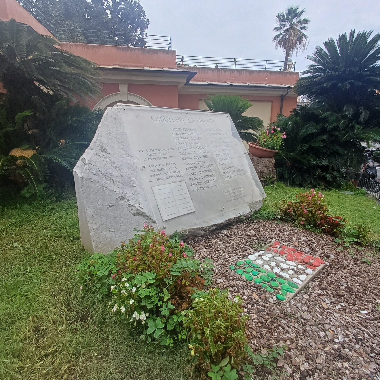 Blocco di pietra grezza con iscrizione, in un giardino con aiuole fiorite e un mosaico tricolore a terra.