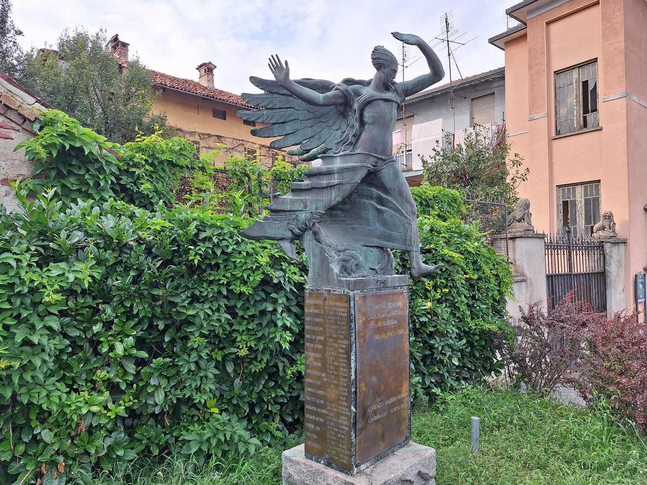 Monumento bronzeo con figura femminile alata su piedistallo con iscrizione, in giardino tra siepi.