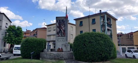 Monumento ai partigiani con bandiera italiana, circondato da verde, in un contesto urbano.