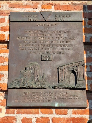 Targa in bronzo su parete di mattoni, con iscrizione e rilievi di un castello e un arco trionfale.