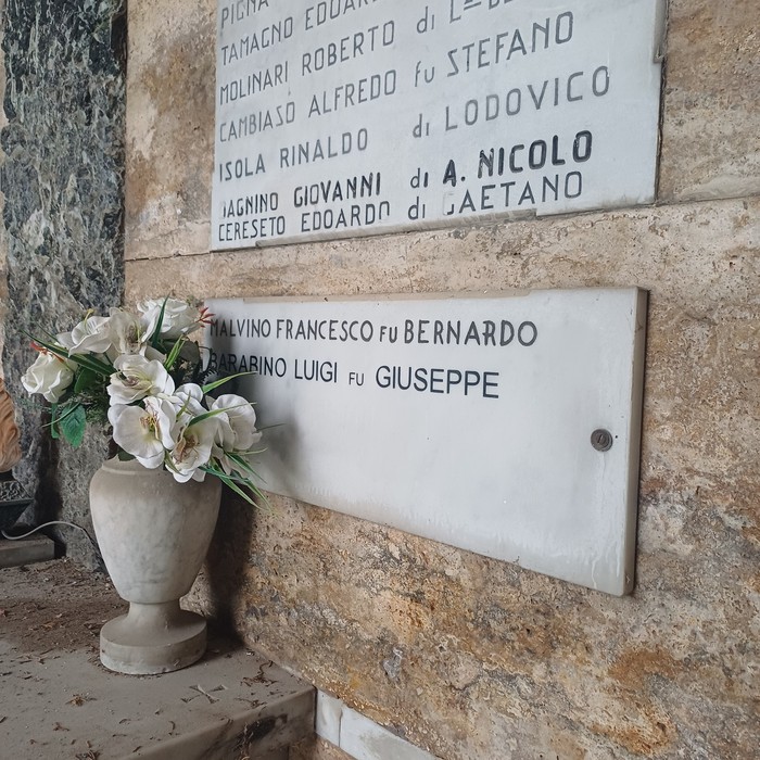 Due lapidi marmoree con elenchi di nomi su parete di pietra, un vaso con fiori bianchi davanti.