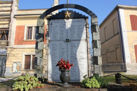 Monumento ai caduti di Vallalta con elenchi di nomi e decorazioni, circondato da piante e architettura storica.