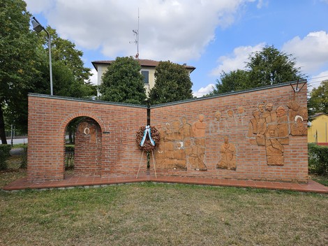 Monumento alla Resistenza con opere in rilievo, circondato da un'area verde e alberi, in un contesto urbano.