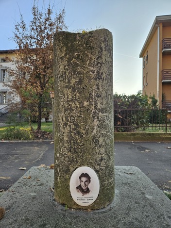Cippo commemorativo con effigie e iscrizioni, circondato da un contesto urbano e vegetazione.