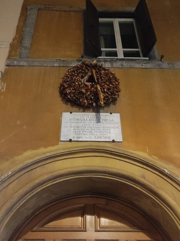 Lapide commemorativa sulla facciata di una chiesa, adornata con una corona di foglie secche.