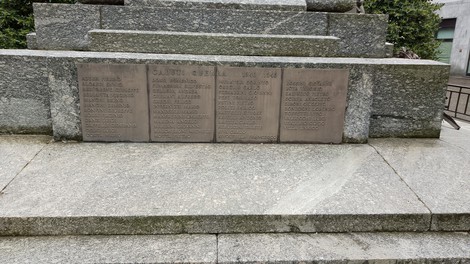 Monumento ai caduti con lastre commemorative incise su pietra, circondato da vegetazione e ambiente urbano.