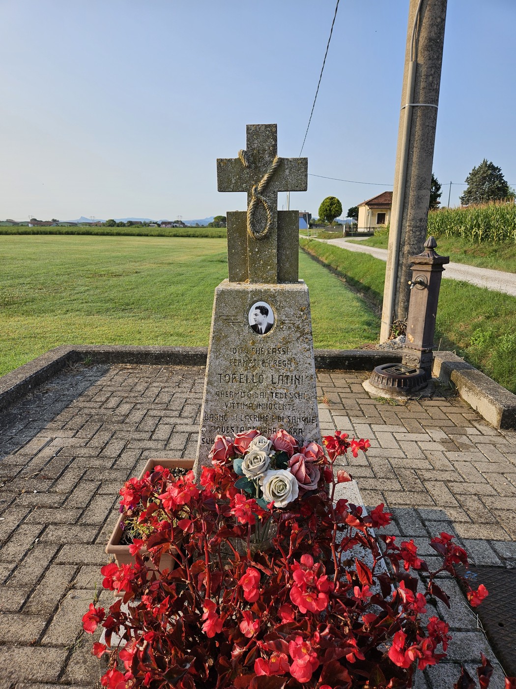 Cippo commemorativo con decorazioni floreali, situato in un paesaggio aperto e sereno.