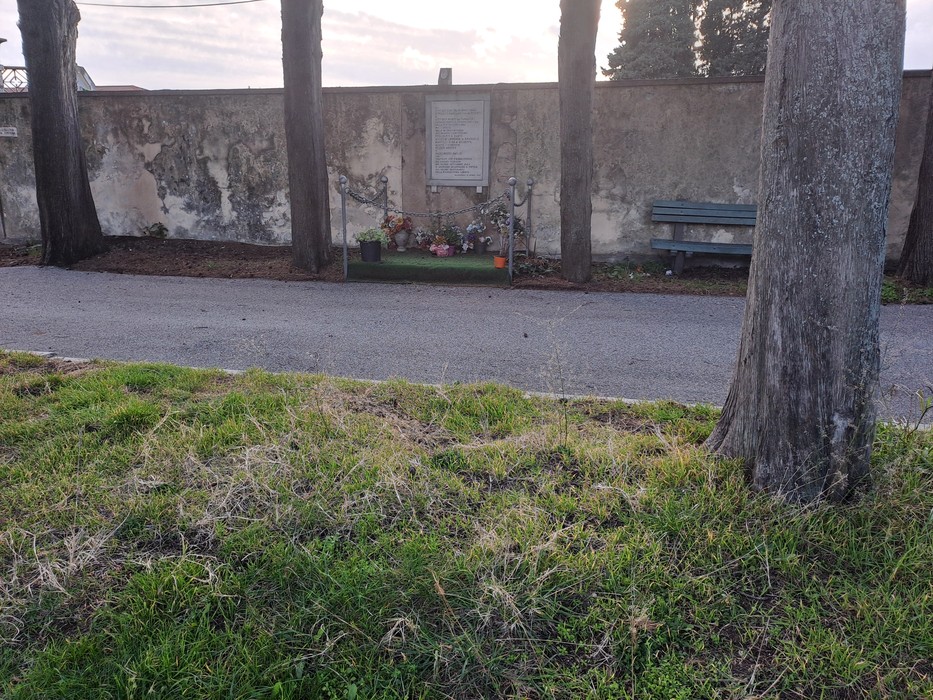 Lapide in un'area verde, circondata da alberi e fiori, con una panchina sullo sfondo.