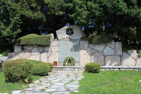 Monumento ai Partigiani al Colletto, circondato da verde e cespugli, con pietre e decorazioni floreali.