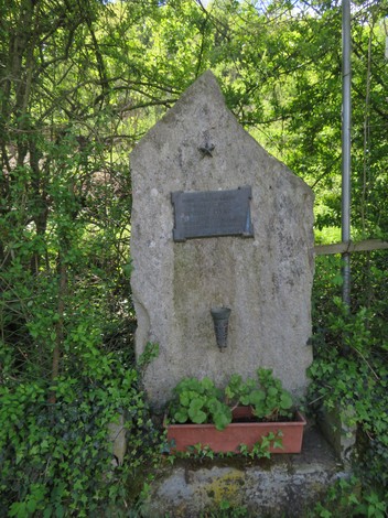 Cippo commemorativo dedicato a Velino, circondato da vegetazione e fiori.